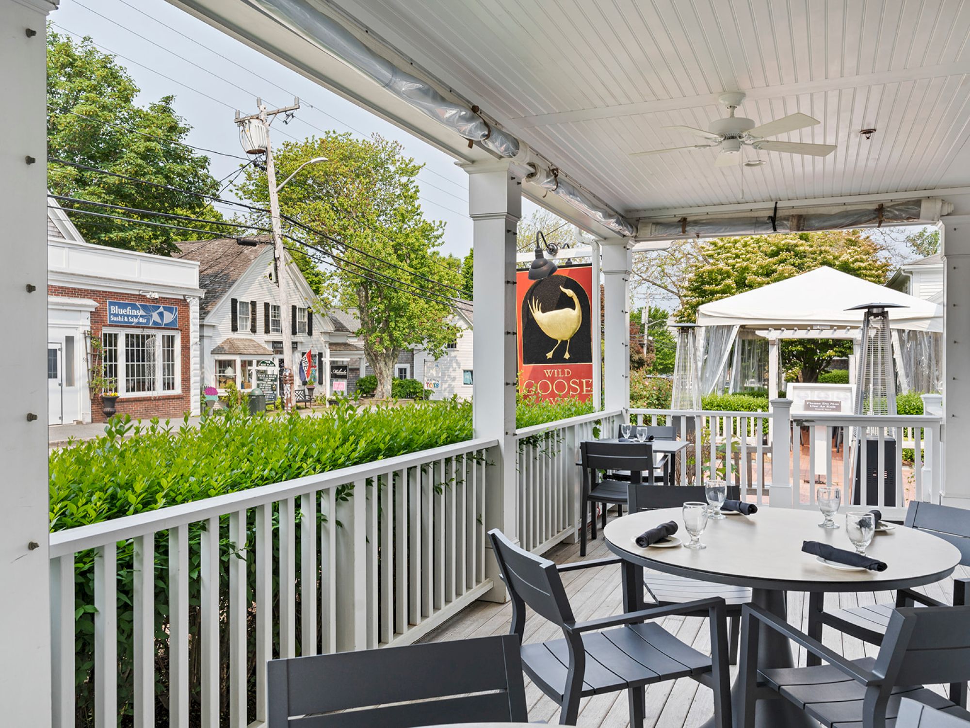 An outdoor patio of a restaurant with a table and chairs, overlooking a street with buildings and greenery, featuring a sign with a goose.