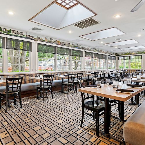 The image shows a bright, spacious restaurant with large windows, wooden tables, black chairs, and a patterned carpet.