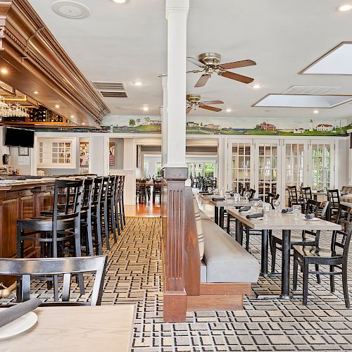 This image shows an empty restaurant dining area with wooden tables, black chairs, a bar with stools, ceiling fans, and large windows.