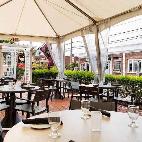 Outdoor dining area with round tables, chairs, glasses, and table settings under a large tent. Green hedges surround the seating area.