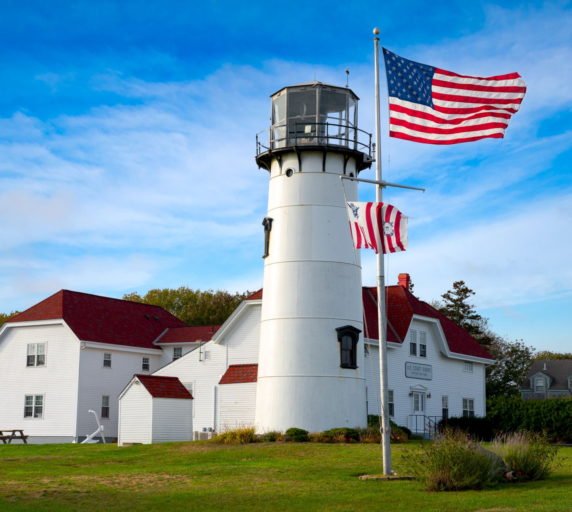 A white lighthouse with a red roof and USA flags on poles, surrounded by green grass under a blue sky with a few clouds is shown in the image.