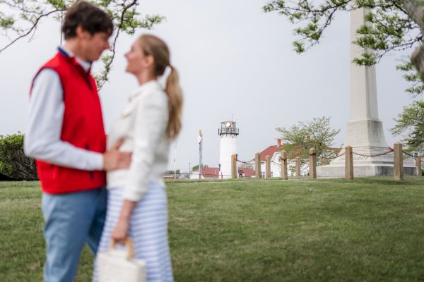 A couple stands closely, facing each other, with a lighthouse and buildings in the background on a grassy area. The scene appears serene and outdoors.