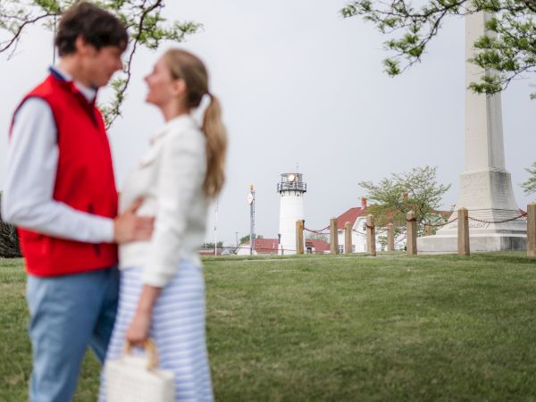 A couple stands closely, facing each other, with a lighthouse and buildings in the background on a grassy area. The scene appears serene and outdoors.