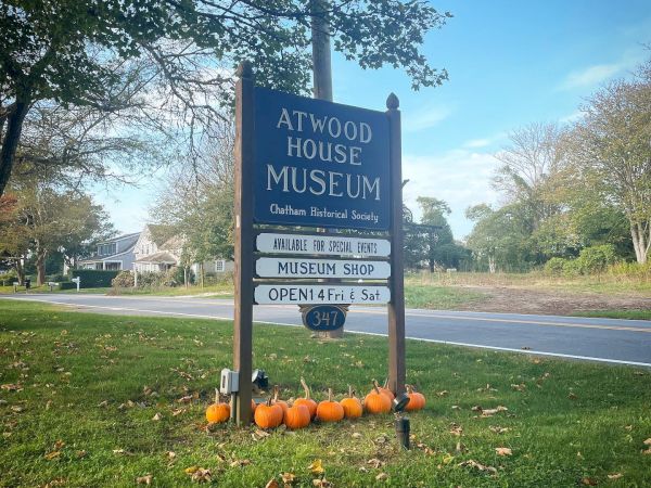The image shows a sign for the Atwood House Museum with details, surrounded by pumpkins, and located next to a road in a green, treed area.