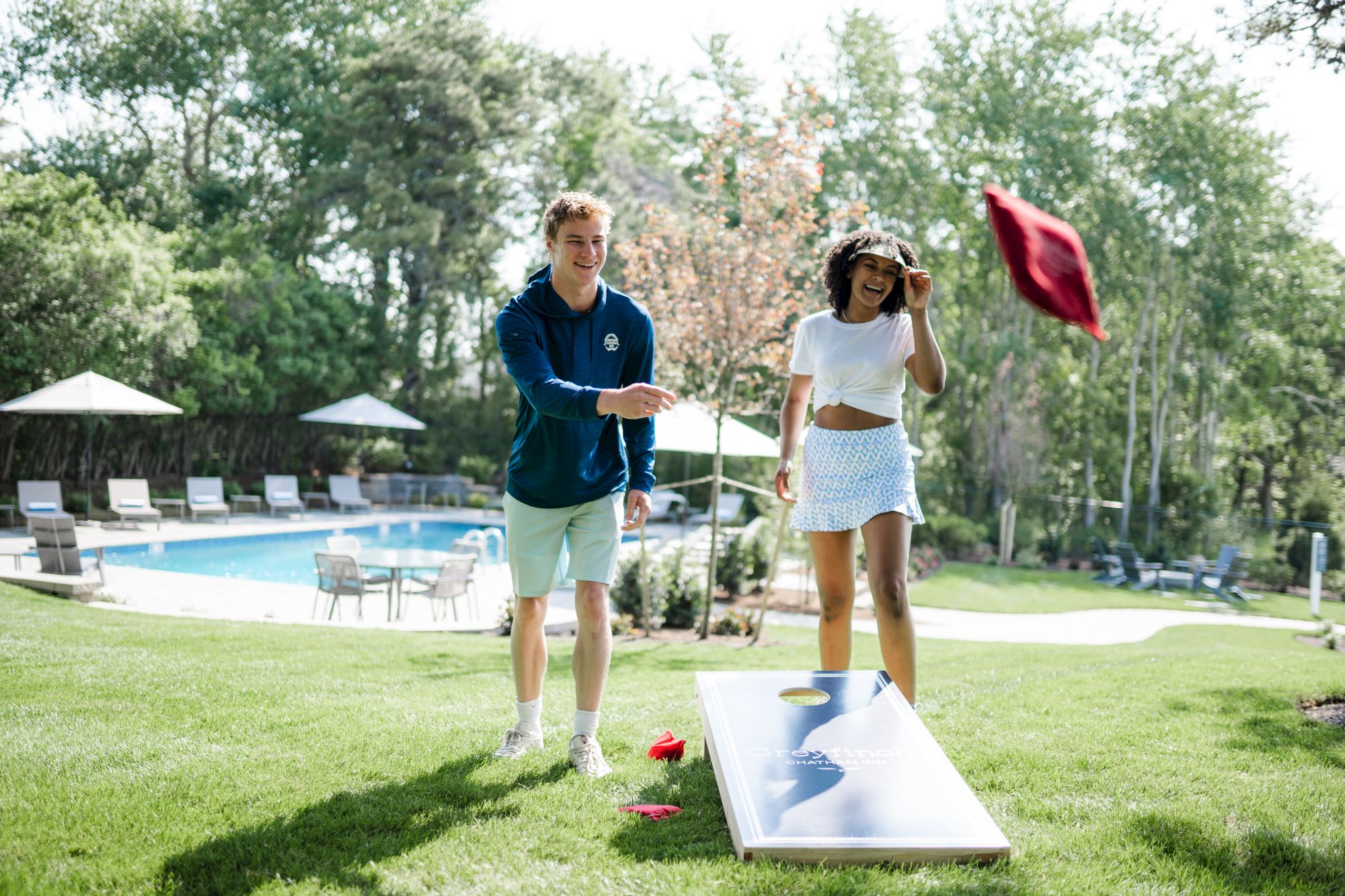 Two people playing cornhole outside near a pool, one throwing a red bag while the other looks on. Trees in the background.