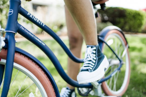 A person riding a blue Priority brand bicycle with red-tinted tires on a grassy area, wearing black and white sneakers.