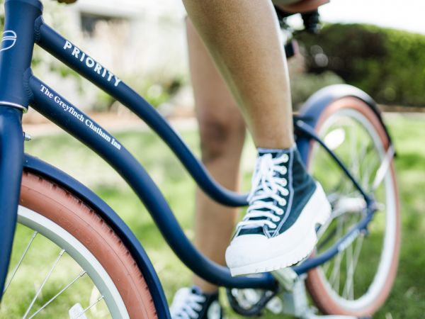 A person riding a blue Priority brand bicycle with red-tinted tires on a grassy area, wearing black and white sneakers.