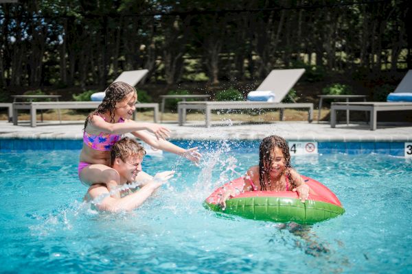 A family is having fun in a swimming pool; one person carries a child on their shoulders, while another child floats on a watermelon-shaped inflatable.