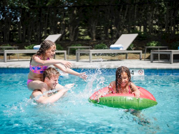 A family is having fun in a swimming pool; one person carries a child on their shoulders, while another child floats on a watermelon-shaped inflatable.