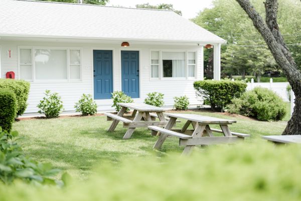 A white building with two blue doors, bushes, and picnic tables in a grassy area, surrounded by trees and greenery, can be seen.