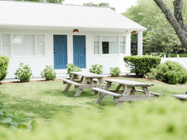 A white building with two blue doors, bushes, and picnic tables in a grassy area, surrounded by trees and greenery, can be seen.