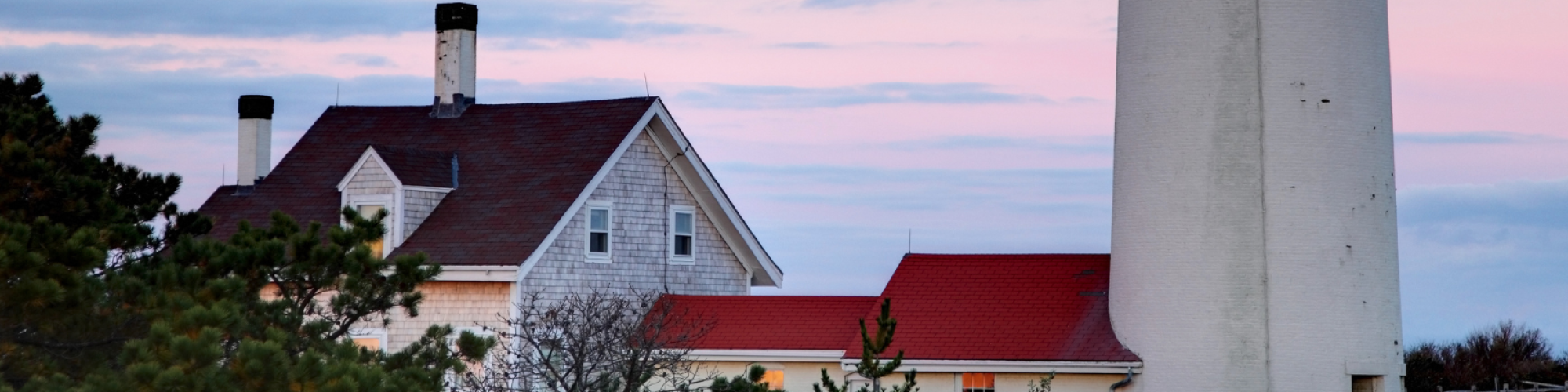 A tall white lighthouse tower stands beside a house with a red roof against a cloudy sky during dusk, surrounded by greenery.