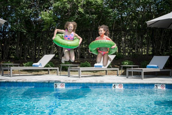 Two children are jumping into a swimming pool with green inflatable rings, with lounge chairs and umbrellas in the background.