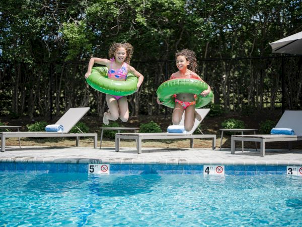 Two children are jumping into a swimming pool with green inflatable rings, with lounge chairs and umbrellas in the background.