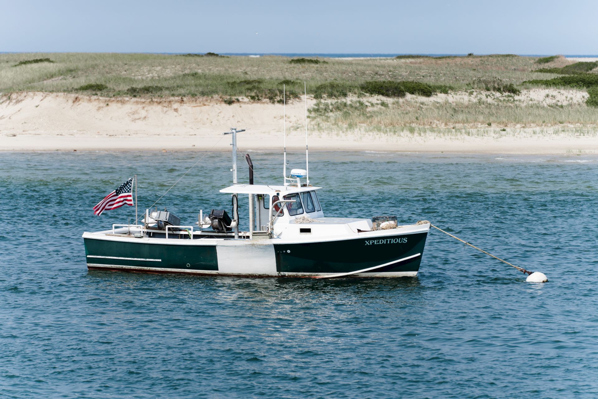 A boat named "MightyMax" is anchored in calm waters near a sandy beach and grassy shore, flying an American flag at the stern.