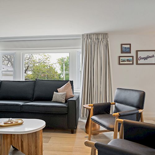 A cozy living room with a black sectional sofa, two black armchairs, a white coffee table, wall art, and large windows with curtains ending the sentence.