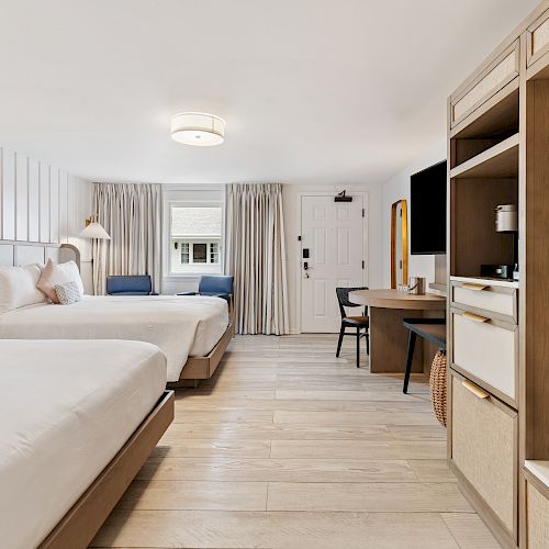 A modern hotel room with two beds, a desk, a flat-screen TV, a wooden floor, light curtains, and lamps on the bedside tables.