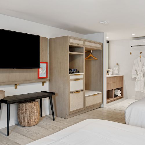 A modern hotel room features two beds, a wall-mounted TV, a small table with a wicker stool, an open wardrobe, and a bathroom area with bathrobes.