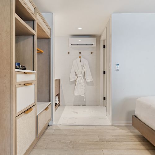 This image shows a modern, minimalistic hotel room with a wooden wardrobe, a white bathrobe hanging on the wall, and a bed. The room is well-lit.