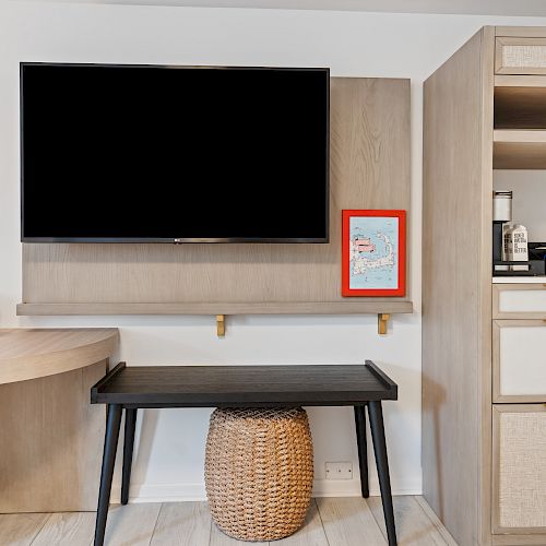 The image shows a modern room with a TV mounted on the wall, a small black table with a wicker basket underneath, and a coffee station in the cabinet.