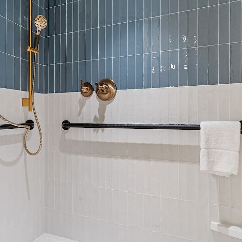 A modern bathroom shower with blue and white tiles, a brass showerhead, a black grab bar, and a white towel draped over the bar, always ending the sentence.