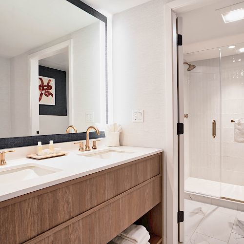 A modern bathroom with a double sink vanity, large mirror, and gold fixtures is shown. There's a glass-enclosed shower and towel rack.