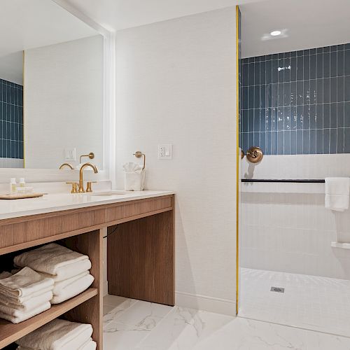 A modern bathroom with a vanity area, mirror, sink, towels, and a walk-in shower with blue tiles and a grab bar, designed for accessibility.