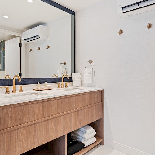 A modern bathroom with a large mirror, wooden vanity, two sinks with gold fixtures, towels, and an air conditioner on the wall.