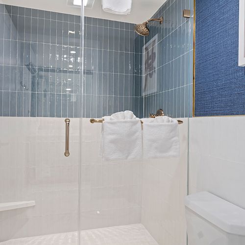 The image shows a modern bathroom with a glass-enclosed shower, blue and white tiles, two hanging towels, and a white toilet.