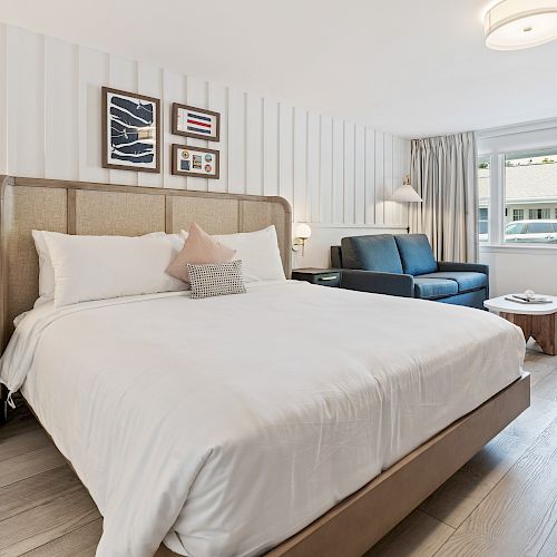 A modern bedroom with a large bed, white linens, artwork on the wall, a nightstand, a sofa, a coffee table, and a window with curtains.