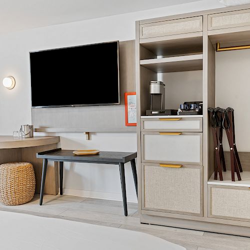 The image shows a modern hotel room with a TV, table, chair, lamp, and an open wardrobe with hangers, a safe box, and a coffee maker on top.