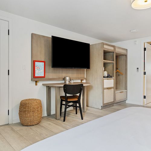 A hotel room with a TV, desk, chair, basket, large mirror, and an adjoining bathroom with modern design and neutral tones.