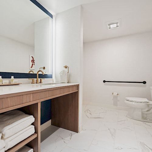 A modern bathroom featuring a large mirror, wooden vanity with towels, marble floor, toilet with grab bars, and wall art.