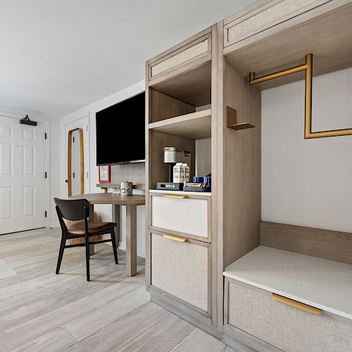 A modern hotel room with a desk, chair, TV, and an open closet featuring hangers and shelves. The space is neatly organized, bright, and welcoming.