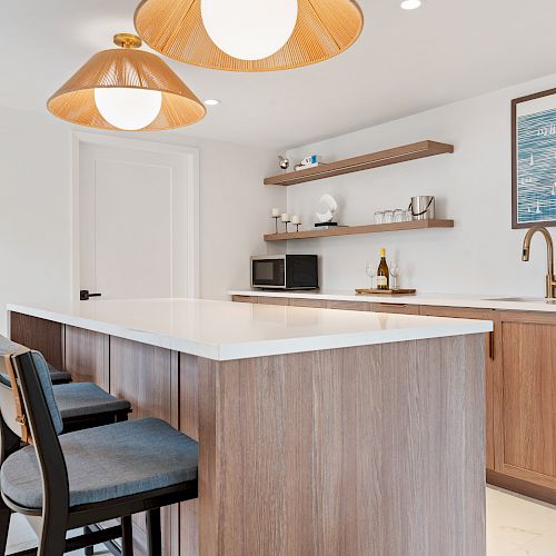 A modern kitchen with a central island, three bar stools, pendant lights, open shelves with dishes, a microwave, a sink, and a dishwasher.