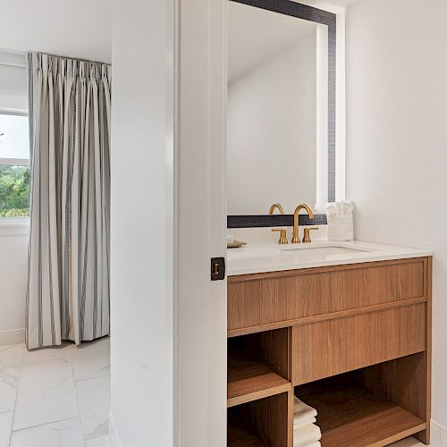 The image shows a modern bathroom with a wooden vanity, gold faucet, a mirror, folded towels below, and a window with curtains.