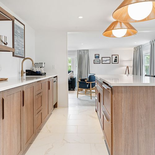 A modern kitchen featuring wooden cabinets, a wine bottle, glasses, and hanging lamps, leading to a dining area with wall art and large windows.