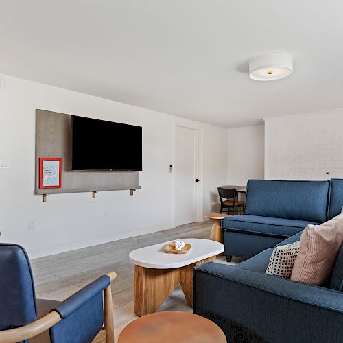 A modern living room with blue sofas, a wall-mounted TV, a small table, and minimal decor. The kitchen area is visible in the background.