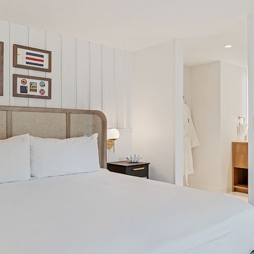 A modern, minimalistic hotel room with a large bed, artwork, and white walls. The bathroom is visible, featuring white robes hanging on the wall.