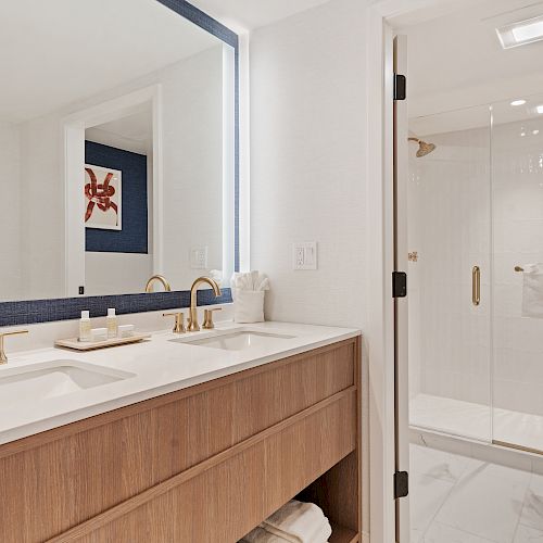 Modern bathroom with a double sink, wooden vanity, large mirror, and glass-enclosed shower with towels on the door.