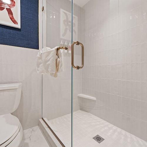 This image shows a bathroom with a toilet, glass shower, white tiles, a towel hanging, and framed art on a dark blue wall ending the sentence.