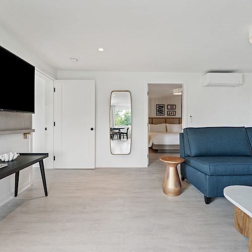 A modern living room with a large TV, blue sectional couch, coffee table, console table, and a wall mirror. Bedroom visible through an open doorway.