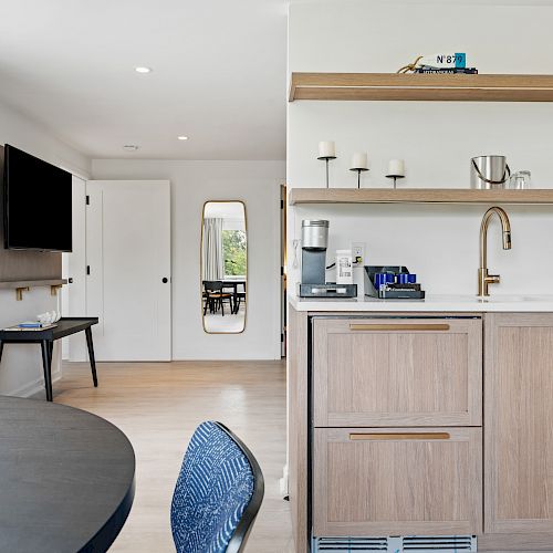 A modern kitchen and living area with a coffee maker, microwave, and open shelving. There is a round table and a wall-mounted TV seen.