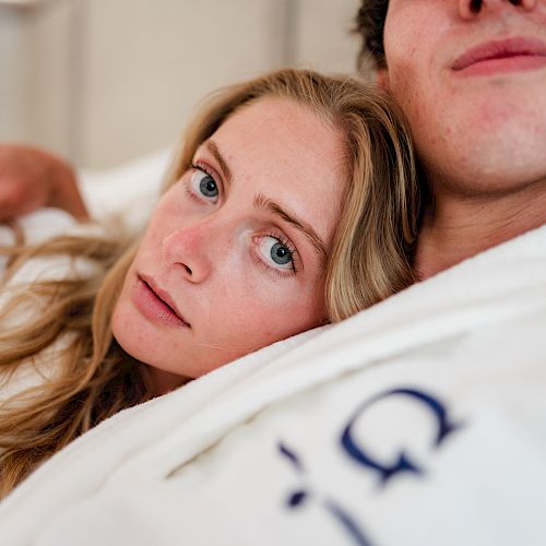 A woman rests her head on a man's chest, both wearing white robes and lying closely together, creating an intimate and relaxed setting.