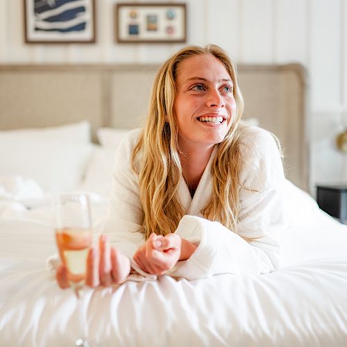 A person in a white robe is lying on a bed, smiling and holding a glass of wine in a cozy room with framed pictures on the wall.