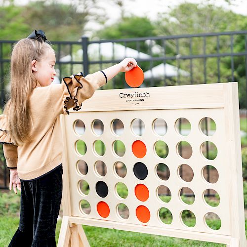 A child is playing a game of giant Connect Four, placing an orange disc into the wooden game board in an outdoor setting, near a fence.