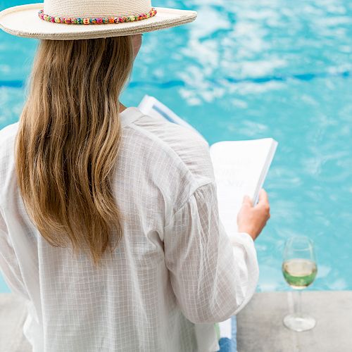 A person with long hair is sitting by a pool, wearing a hat and holding a book, with a glass of wine next to them.