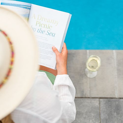 A person wearing a white hat reads a book titled "A Dreamy Picnic by the Sea" next to a pool, with a glass of white wine on the ground.