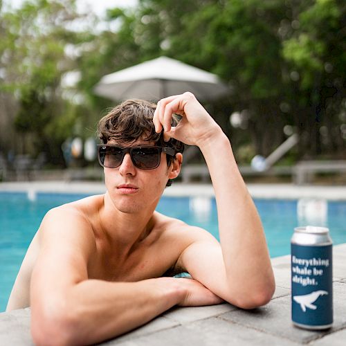 A person wearing sunglasses is lounging by a pool, with a beverage can on the ledge next to them and greenery in the background.