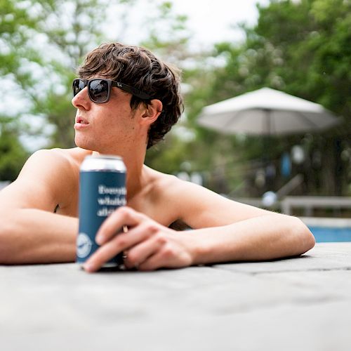 A person lounges by a pool, holding a drink can, and wearing sunglasses, with greenery and an umbrella in the background.